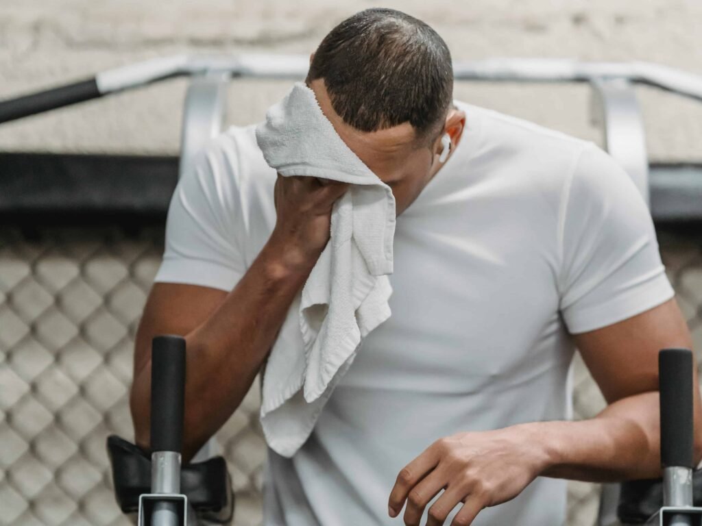 A man in a white shirt wipes sweat from his face with a towel while exercising on gym equipment.